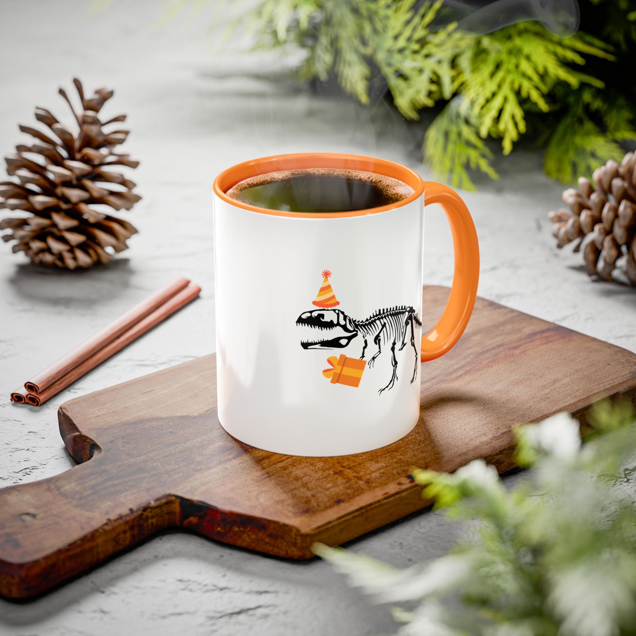 White mug with orange handle featuring a dinosaur skeleton design on a wooden board with pinecones and cinnamon sticks.