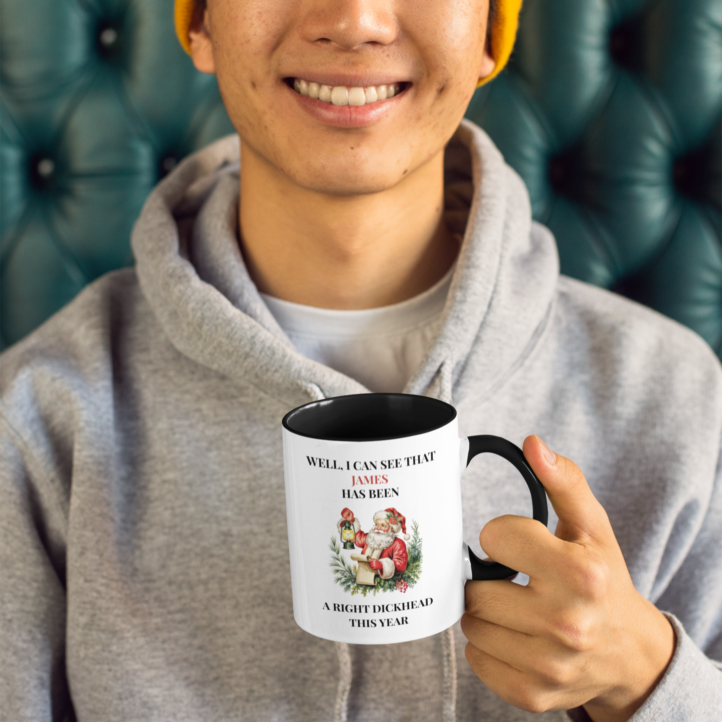 Person holding a mug with text and graphics, wearing a yellow beanie and gray hoodie, sitting on a green leather couch.