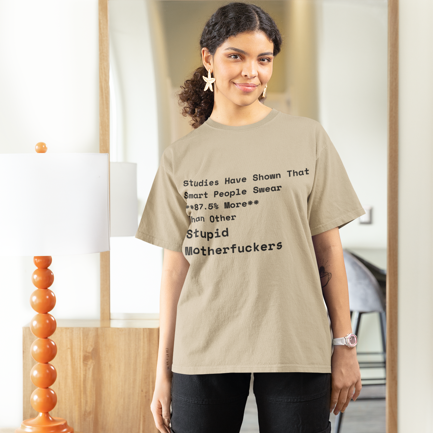 Woman wearing a sand colored t-shirt with text in a room with a mirror and decor.