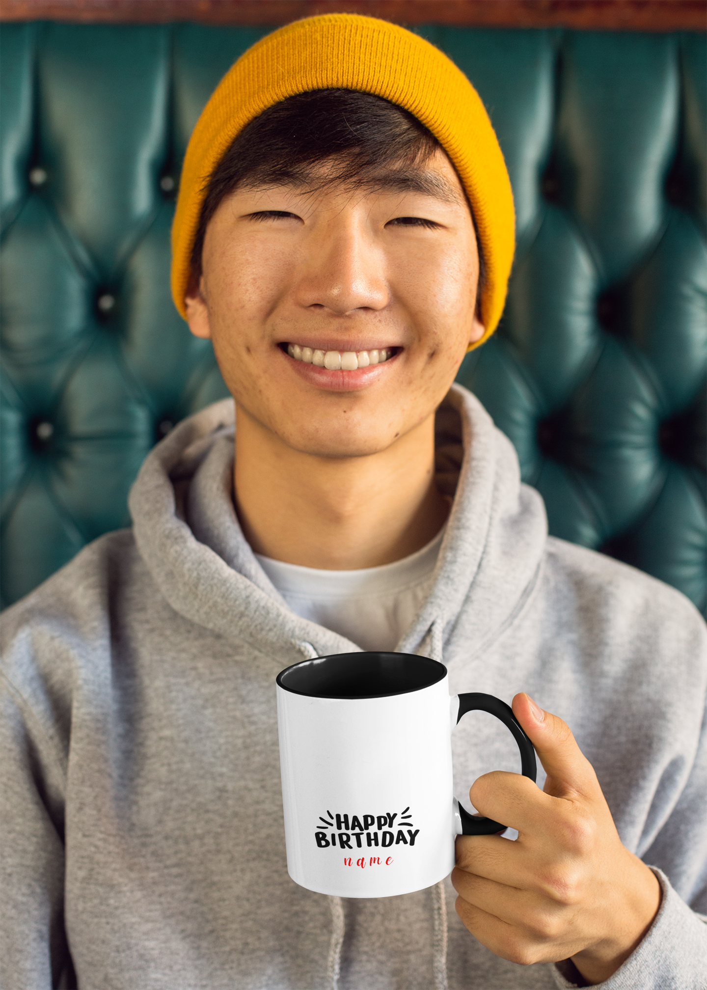 Person wearing a yellow beanie and gray hoodie holding a mug with 'Happy Birthday' text.
