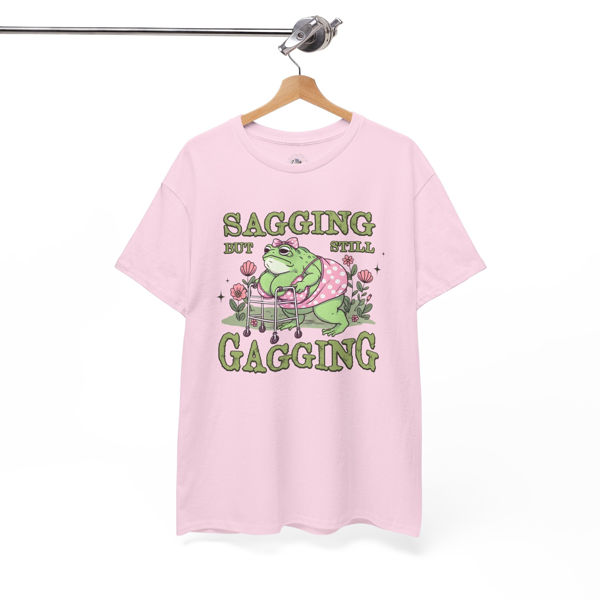 Pink t-shirt with a graphic of a frog and text 'Sagging but still gagging' on a hanger against a white background.