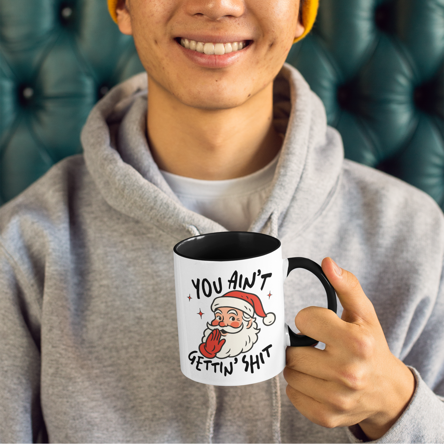 Person holding a mug with a humorous design, wearing a yellow beanie and gray hoodie, sitting on a green leather couch.
