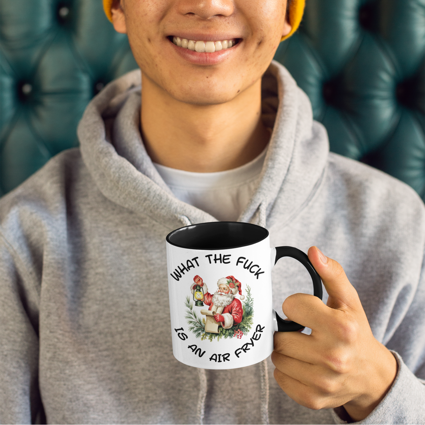 Person holding a sarcastic christmas gift mug and graphics, wearing a yellow beanie and gray hoodie, sitting on a green leather couch.